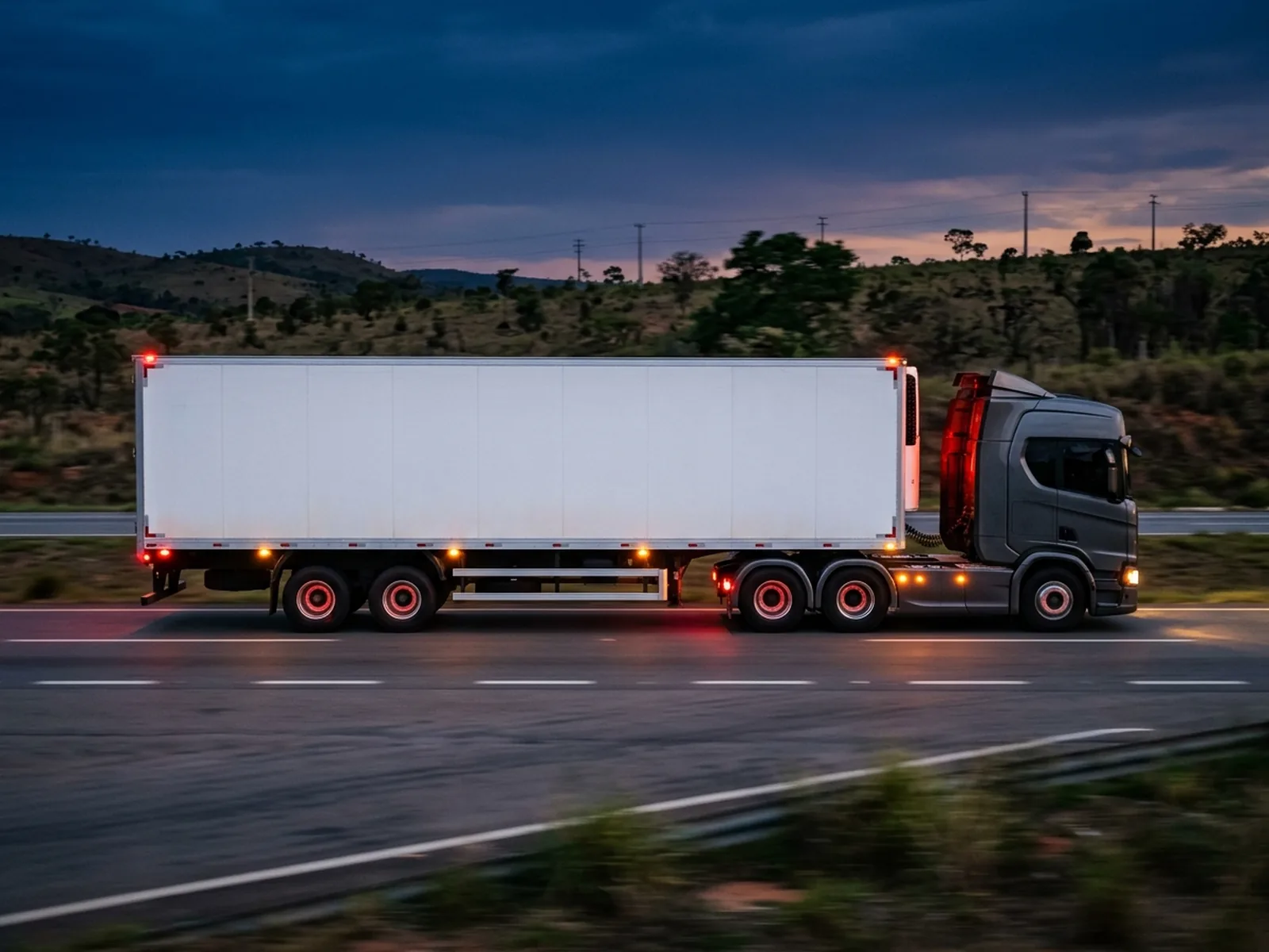 Carreta baú branca com cavalo grafite em movimento numa rodovia, vista lateral ao anoitecer