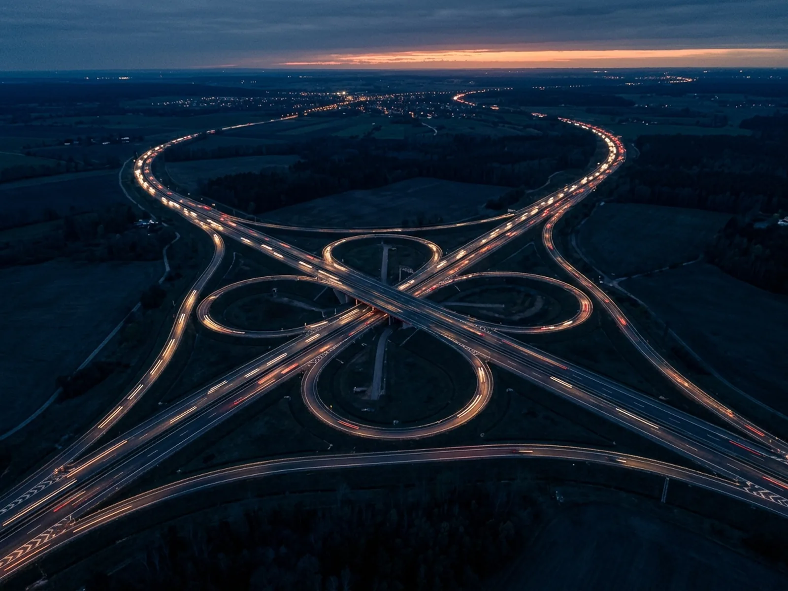 Vista aérea de um trevo rodoviário ao anoitecer, com rastros de luz dos veículos
